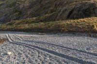 a lone sheep stands in the middle of a gravel field near the water and mountains