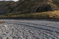 a lone sheep stands in the middle of a gravel field near the water and mountains