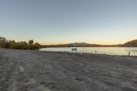 an empty dirt area with benches on the shore of water near a lake with mountains and trees in the distance