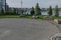 a park near the water with rocks on the road and houses on the other side