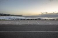 a empty parking lot next to the ocean with waves in the background while someone walks down the road