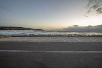 a empty parking lot next to the ocean with waves in the background while someone walks down the road