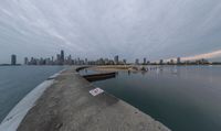 a cloudy view shows the water below a pier on lakefront with buildings in the distance