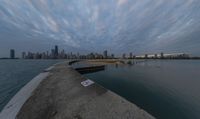 a cloudy view shows the water below a pier on lakefront with buildings in the distance