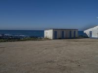 two old white shed houses on the side of a beach with water in background and a jet plane overhead