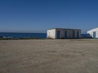 two old white shed houses on the side of a beach with water in background and a jet plane overhead