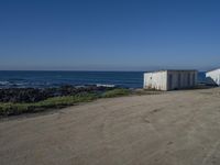 two old white shed houses on the side of a beach with water in background and a jet plane overhead