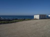 two old white shed houses on the side of a beach with water in background and a jet plane overhead