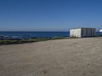 two old white shed houses on the side of a beach with water in background and a jet plane overhead