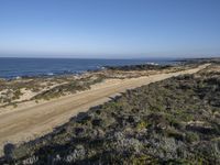 a dirt road runs near the water in front of some sand dunes and shrubs, with two people walking on it