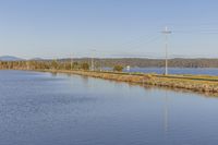 the train is traveling along side of the road by the water and utility lines across the river