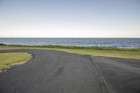 the asphalt road runs next to the sea and grassy area at dusk and it appears to be winding