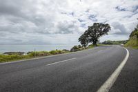 an empty road with a beach in the distance below the clouds and water along side a rocky cliff and tree