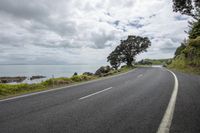 an empty road with a beach in the distance below the clouds and water along side a rocky cliff and tree