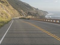 a motorcycle is riding on the road by the ocean and cliffs in the background with waves on the sea