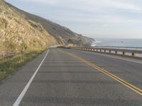 a motorcycle is riding on the road by the ocean and cliffs in the background with waves on the sea