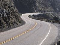 an empty long straight highway passes by on a cloudy day under a mountain range with large rocks and cliffs