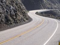 an empty long straight highway passes by on a cloudy day under a mountain range with large rocks and cliffs