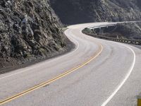 an empty long straight highway passes by on a cloudy day under a mountain range with large rocks and cliffs