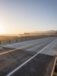 an empty road near the coast is lit by the setting sun with a mountain view