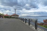 a street with fence and houses near the water shore on a cloudy day, some cars and buses parked in parking