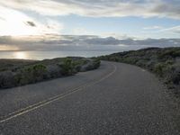 the road winds through the desert near the water with clouds at sunset above it and a few shrubs below