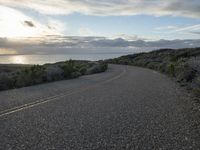 the road winds through the desert near the water with clouds at sunset above it and a few shrubs below