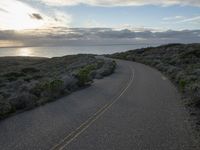 the road winds through the desert near the water with clouds at sunset above it and a few shrubs below