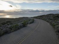 the road winds through the desert near the water with clouds at sunset above it and a few shrubs below