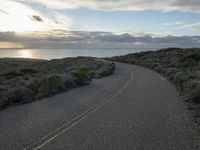 the road winds through the desert near the water with clouds at sunset above it and a few shrubs below