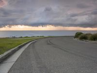 a paved road leads to the ocean with clouds overhead and a bench in the corner