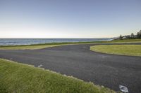 a empty road that runs along the shore with the water in the distance near a grassy field