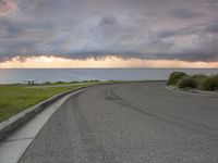 a street view of the ocean and clouds above a beach with a bench in the grass