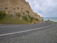 a curve on a road near the water and a cliff face in the background with waves rolling in