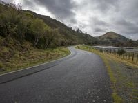 an empty road stretches along a forested hillside and water is in the distance under gray, grey clouds