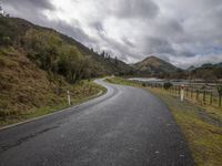 an empty road stretches along a forested hillside and water is in the distance under gray, grey clouds