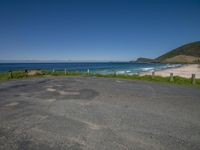 the empty road is next to the grassy hill near the ocean and sand beach in the distance