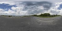 a fish eye lens that has clouds overhead and a blue sky behind it and a road, trees, a lake, a dirt road, and green grassy hill