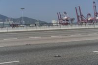 a roadway with a fence, a traffic light and cranes in the background in a view of a harbor