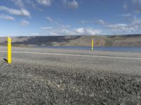 a yellow post at the corner of a road by a body of water with hills and clouds in the distance