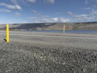 a yellow post at the corner of a road by a body of water with hills and clouds in the distance