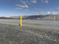 a yellow post at the corner of a road by a body of water with hills and clouds in the distance