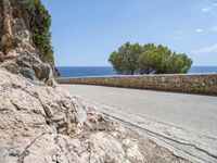 a long road on a rocky hill near water and trees with blue sky and clouds