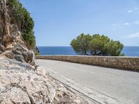 a long road on a rocky hill near water and trees with blue sky and clouds