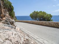 a long road on a rocky hill near water and trees with blue sky and clouds