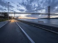 an empty road with a bridge and the ocean in the background at sunset with a sky full of clouds