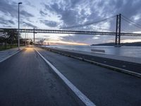 an empty road with a bridge and the ocean in the background at sunset with a sky full of clouds