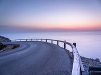 a curved road over the ocean with a beautiful sunset behind it, and the ocean in the distance