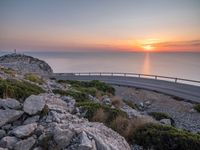 rocks along a road overlooking a large body of water at sunset time with cars driving on the track