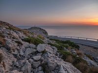 rocks along a road overlooking a large body of water at sunset time with cars driving on the track